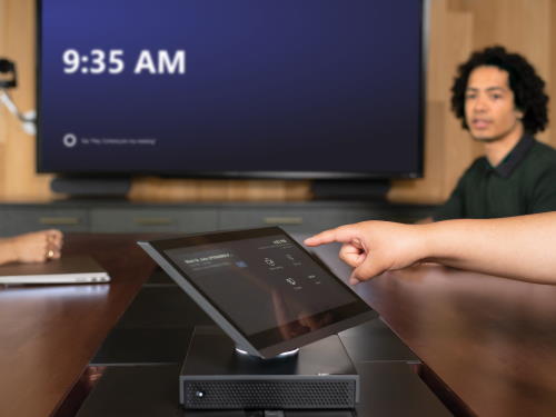 A user taps a Teams Rooms console, with a display in the background A user taps a Teams Rooms console, with a display in the background.